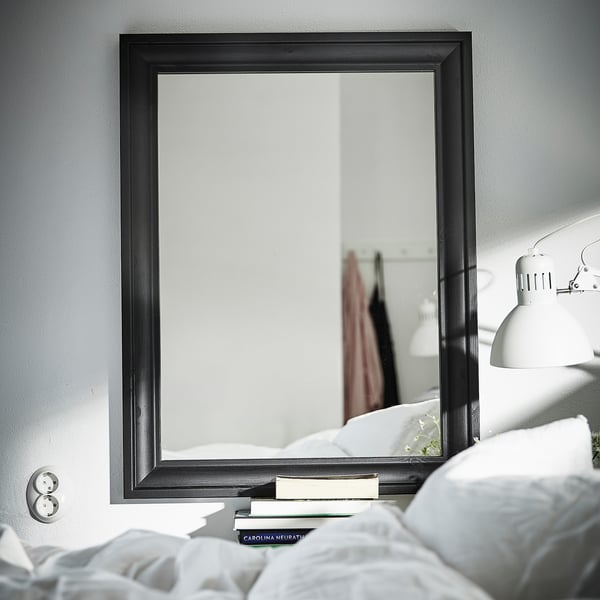 Zwarte TOFTBYN-spiegel op grijze muur, reflecterende slaapkamer met bed, boeken en lamp.