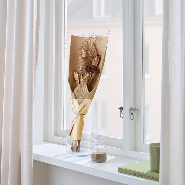 Gedroogde bloemen in bruin papier op vensterplank met zandloper en boek.