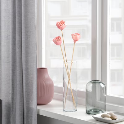 Drie roze bloemen in een heldere glazen vaas op een vensterbank, geflankeerd door twee decoratieve vazen.