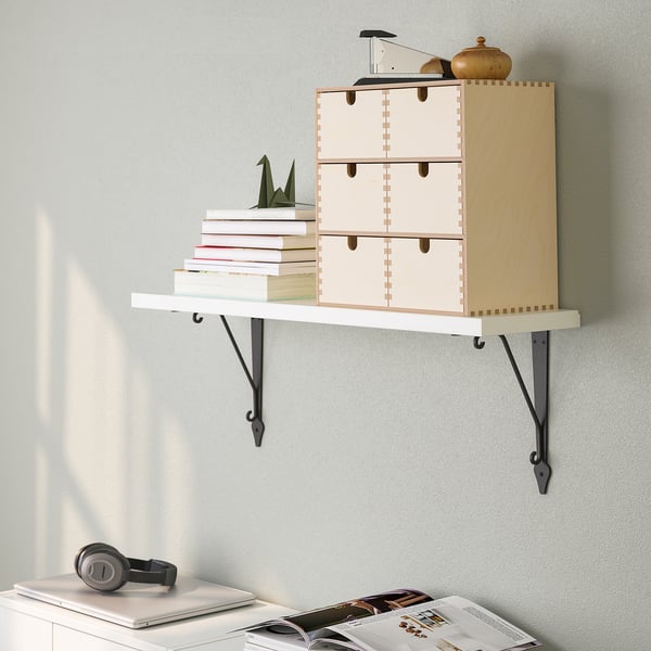 Een aan de muur gemonteerde plank met lades met boeken en een decoratief item. Hieronder een bureau met koptelefoons, een laptop en tijdschriften.
