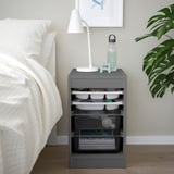 Grijze TROFAST opberger bij bed met planken en dienbladen, gegarneerd met lamp, fles en speelgoed.