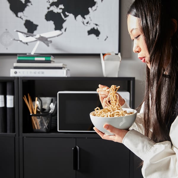Vrouw die noedels uit een kom eet terwijl ze voor een bureau zit met een computer en een boekenplank op de achtergrond.