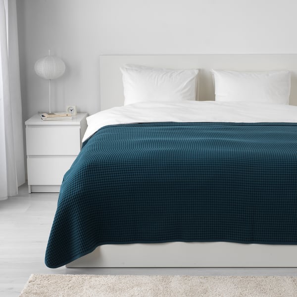 Donkerblauwe VÅRELD katoenen sprei met witte kussens, moderne kamer, klein nachtkastje met lamp en boek.