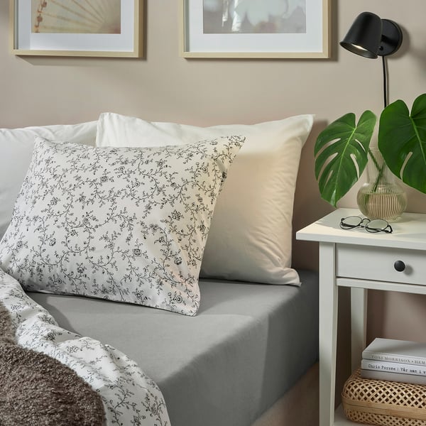 Gezellig bed met VÅRVIAL lakens, bloemenkussen, nachtkastje, lamp, plant en mand met boeken.