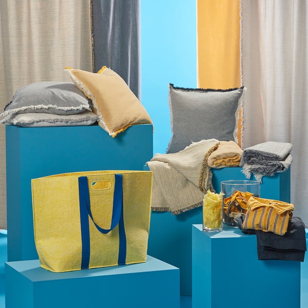 Weergave van de ikeas VÄXELBRUK collectie: gele draagtas met blauwe hengsels, diverse kussens en gevouwen stoffen op blauwe standaards met levendige gordijnen.