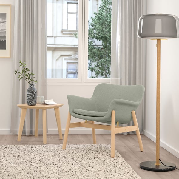 Minimalistische woonkamer met lichtgrijze VEDBO fauteuil, houten bijzettafel, hoge lamp en groot raam.