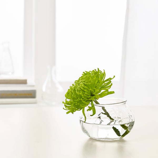 Kleine glazen vaas, VILJESTARK, bevat één groene bloem. Strak, rond ontwerp, zit op een witte tafel.