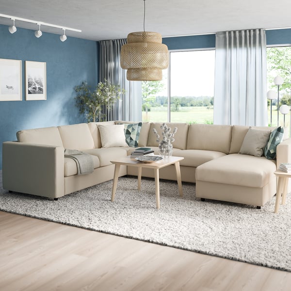 Moderne woonkamer met grote beige sectionele bank, chaise longue, salontafel, blauwe accentmuur, vloerkleed en ramen met gordijnen.
