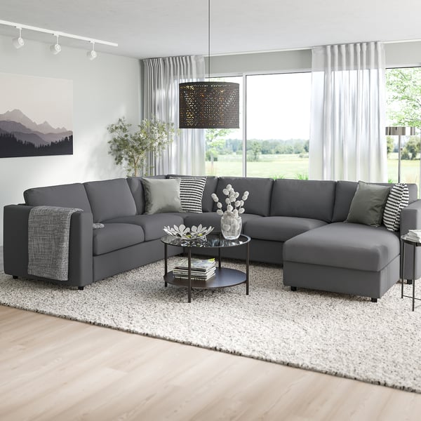 Moderne 3-delige grijze sectionele bank met chaise longue ; ruim, converteerbaar om te slapen, omgeven door grote ramen en decor, op een licht kleed.