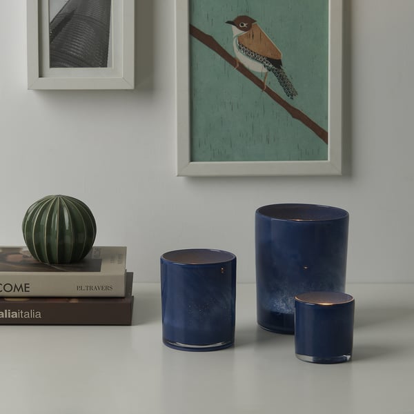 Drie blauwe glazen kandelaars met getextureerde patronen op tafel, naast groene glazen bol, onder ingelijste kunst.