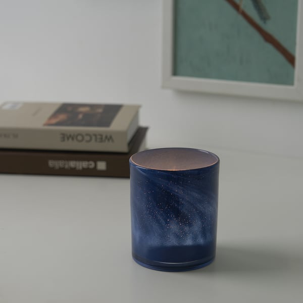 Een blauwe, gespikkelde kandelaar staat op een witte tafel, met een brandende kaars erin, die een warme gloed werpt. Daarachter zijn een boek en een lijst zichtbaar.