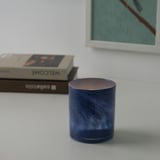 Een blauwe, gespikkelde kandelaar staat op een witte tafel, met een brandende kaars erin, die een warme gloed werpt. Daarachter zijn een boek en een lijst zichtbaar.