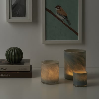 Drie VINDSTILLA kaarsen van verschillende groottes op tafel met boeken, aangestoken en warm gloeiend. Kunst op de achtergrond.