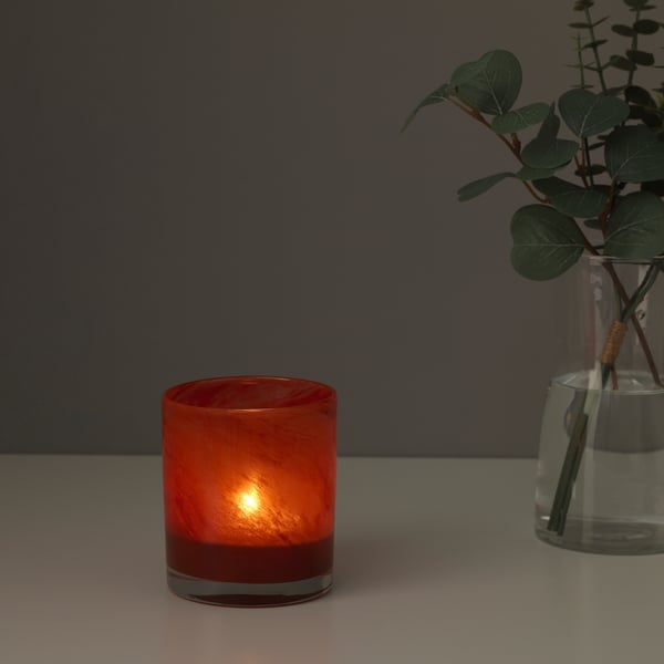 Een kleine, rode, glazen kandelaar gloeit zachtjes op een tafel en werpt een warm licht. Ernaast staat een glazen vaas met groene bladeren.