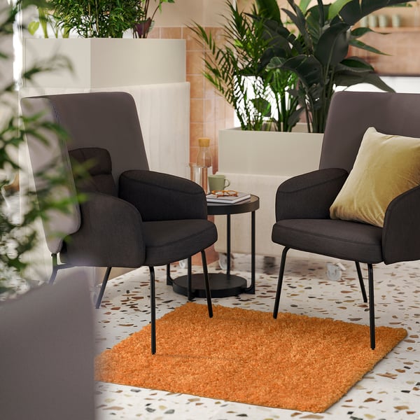 Two grey ÄRENDE chairs with black legs and yellow pillows on an orange rug, surrounded by plants and a black side table with a glass mug.