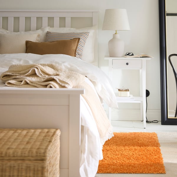 A cosy bedroom with a white bed, neutral pillows, and an orange ÄRENDE rug. A bedside table has a lamp and items, with a black chair at the edge.