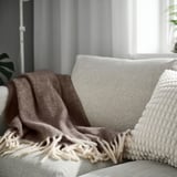 ALLFRAKEN grey wool blanket with fringes on beige sofa, decorates large room.