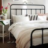 Bedroom with black metal bed, green-white duvet, striped pillows, and beige blanket. White bedside table with pink vase, books, and slippers on rug.