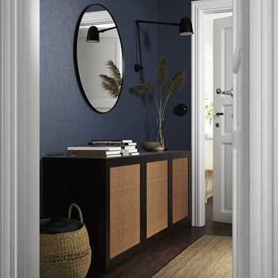 A modern hallway: BESTÅ cabinet, black & wicker, rectangular, with books & flowers. Against blue wall, under round mirror, beside a white door.
