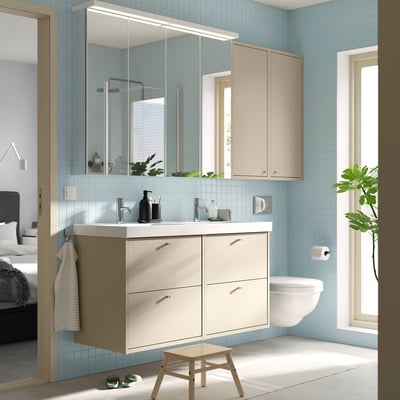 Modern beige bathroom cabinet with dual basins, large mirror, wall-mounted toilet, small stool, and green plant by window.