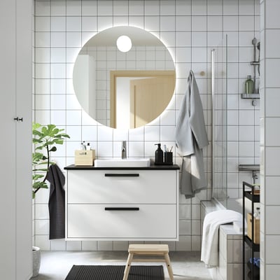 Modern bathroom with white HAVBÄCK vanity, round mirror, grey towels, plant, and stool.