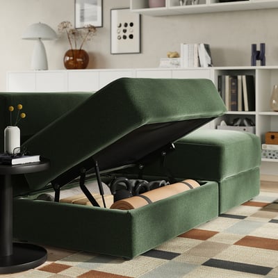Green convertible sofa with open storage compartment revealing pillows, set in a cosy room with shelves and decor.