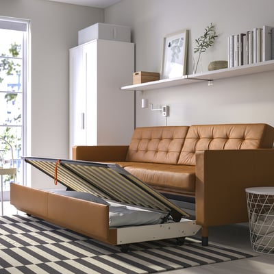 A tan-colored LANDSKRONA sofa-bed in a room. Its large, with storage and a mattress under the seat, positioned against a wall with shelves.
