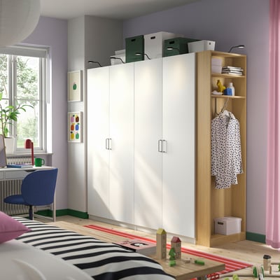 Modern white PAX wardrobe with wooden end unit. Spacious, minimalist design, multiple compartments. Bright, airy room with desk and window.