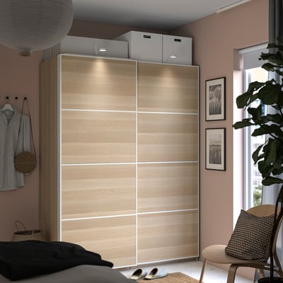 Modern bedroom with a large light wooden sliding door wardrobe, featuring integrated lighting, a cosy bed, and lush green plants.