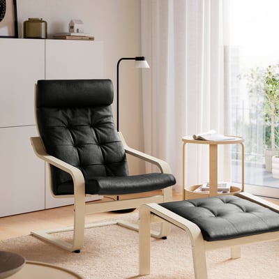 Modern living room with black leather POÄNG chair, cushions, footstool, side table, and floor lamp by window.