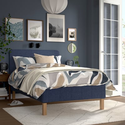 Modern bedroom with blue headboard bed, dark blue wall, abstract duvet, artwork, large window, cream rug, plant, and bedside table with flowers.
