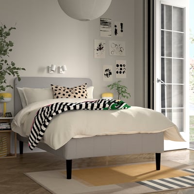 Modern bedroom with RÅHOLMAR grey bed, striped blanket, yellow lamp, green plant, abstract art, and patterned rug. Natural light brightens the room.