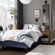 Modern bedroom: grey bed with leaf print duvet, wooden bedside table, grey walls, large plant, and hanging clock.