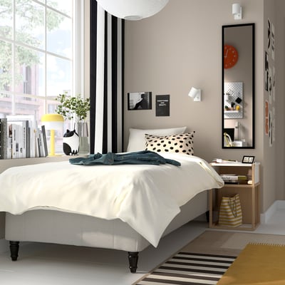 Modern bedroom with råholm bed, polka-dot pillow, blue jacket, wooden bedside table, yellow lamp, books, decor, striped rug, natural light.