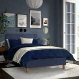 Modern blue bed in room with dark walls. Large white door, art on walls, layered bedding.