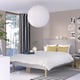 Modern minimalist bedroom: large bed, white linen, mustard pillows, black curtains, round white pendant, striped rug, art wall, plants.
