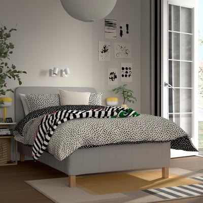 Modern bedroom with grey bed, polka-dot & striped bedding, plant, abstract art, side table with lamp, plant, book, frame, large window, striped rug.