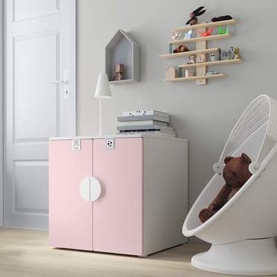 A pastel pink childrens storage cabinet with a grey top, white adjustable legs, and soft-close hinges next to a white rocker with a teddy bear.