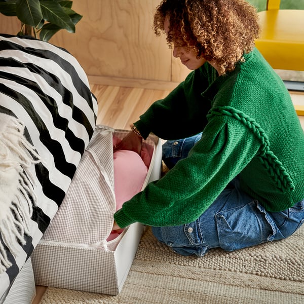 Person sitting, arranging pillows on a bed. The bed has a striped pillow and a basket with cushions.