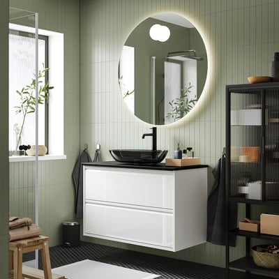 A modern bathroom with a wall-mounted white ÄNGSJÖN wash-stand featuring minimal design, a dark glass vessel sink, round mirror, and black accents.