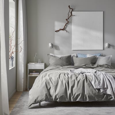 Modern minimalist bedroom with large white bed, grey linens, textured wall art, and sleek bedside table. Soft lighting and calming colours.
