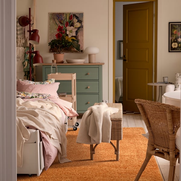 Cosy bedroom with large beige ÄRENDE rug, white bed, green dresser, red lamp, vase, and painting.