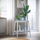 White ALPVIDE plant stand displays large green plants near a window, featuring a steel tray for easy watering and small tools.