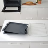 Modern kitchen with black baking trays, cooling rack, and loaf of bread on counter near open oven.