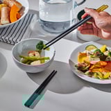 A person holds colourful chopsticks reaching into a bowl of shrimp and vegetable soup. Bowls and plates of food surround them on a white table.