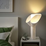 Lit white lamp with flower shade on bedside table. Bed has green/white pillows, glass with cups.