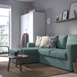 Modern living room with teal barslov sofa-bed and side table. Decorative pillows, grey walls, and natural light from a window. A cosy, inviting space.