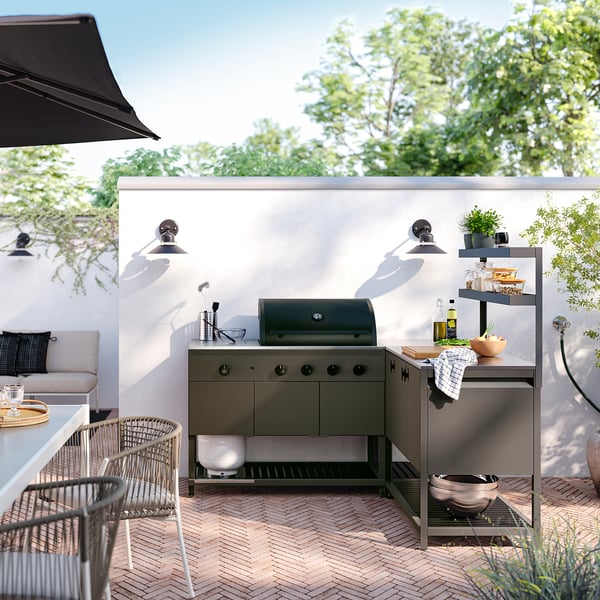 Modern outdoor kitchen with grey metal cabinets, grill, and shelves. Surrounded by patio furniture and greenery.