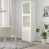 White BESTÅ cabinet with glass doors, displaying dishes, beside desk, chair & plant on rug, window background.