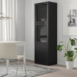 Black BESTÅ cabinet with glass doors, standing tall near a desk & chair, displaying wine glasses & dishes, next to a large leafy plant.